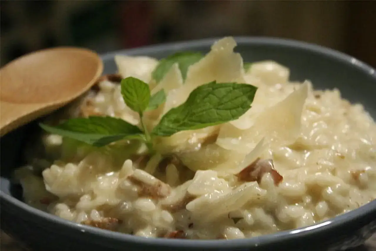 Un cr&eacute;meux risotto aux champignons, garni de copeaux de parmesan et de feuilles de menthe fra&icirc;che. Une cuill&egrave;re en bois est pos&eacute;e &agrave; c&ocirc;t&eacute;.