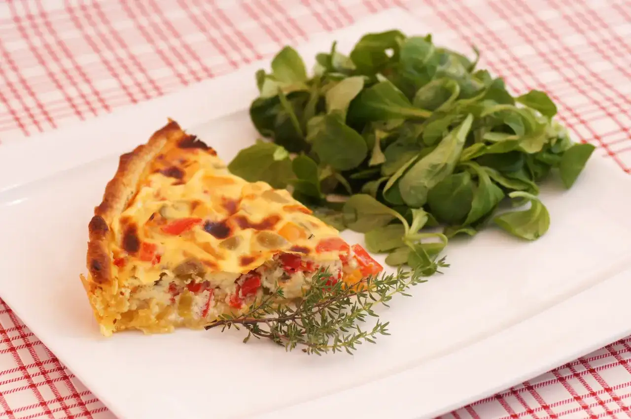 Une part de tarte aux poivrons colorés, garnie d'une branche de thym, accompagnée d'une salade verte fraîche sur une assiette blanche.