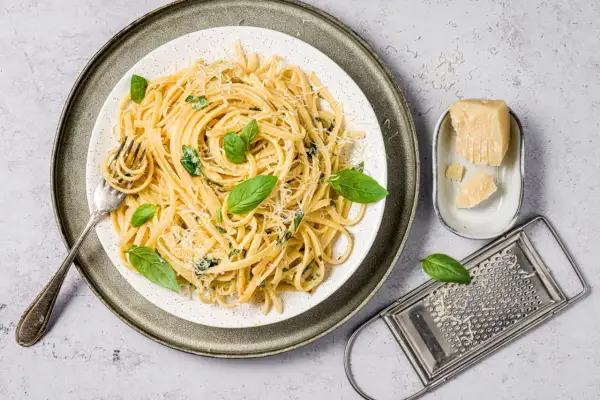 Assiette de pâtes au basilic frais et parmesan râpé, avec une fourchette prête à déguster. Un morceau de parmesan et une râpe complètent la scène.