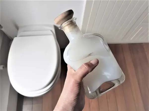Main tenant une bouteille en verre remplie d'un produit wc maison, prêt à nettoyer la cuvette blanche.