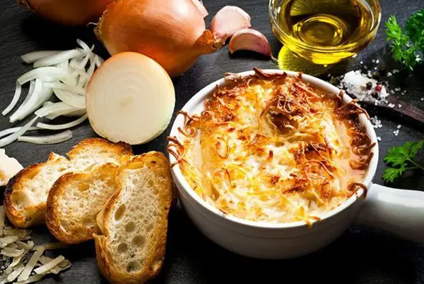Une délicieuse soupe à l'oignon gratinée, servie avec des croûtons croustillants et du fromage fondu. Les ingrédients frais, comme les oignons et l'ail, sont disposés autour du plat.