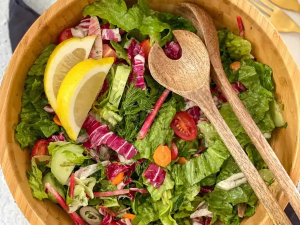 Salade verte fraîche et colorée, parfaite pour une recette légère. Radis, concombre, tomates cerises et citron dans un saladier en bois avec des couverts.