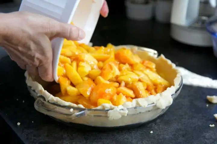 Une main verse des morceaux de pêches en boîte dans une tarte.